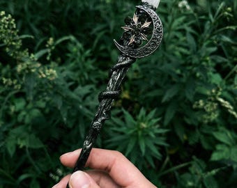 Könnte beinhalten: Ein schwarzer Zauberstab mit Kristallspitze, Mond und Stern-Design. Die Stange hat eine Schlange, die sich um den Schaft windet. Der Zauberstab wird vor einem Hintergrund aus grünem Laub gehalten.