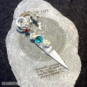 Może przedstawiać: Ozdobny otwieracz do listów ze srebrnym smokiem, białymi różami i niebieskim klejnotem w kształcie oka. Ostrze jest białe, a uchwyt ozdobiony czerwoną perłą. Otwieracz spoczywa na szarej kamiennej płycie z tekstem.