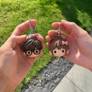 May include: Two character keychains made of clay. One features a figure with brown hair and black glasses attached to a star charm. The other features a figure with brown hair, a pink bow, and pearl-like earrings attached to a crescent moon charm.
