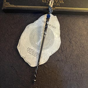 Puede incluir: Una varita m&aacute;gica artesanal con una punta de cristal transparente, una rosa azul y detalles plateados y azules. La varita descansa sobre una piedra con las palabras "Tree of Life" y una caja decorativa. La varita mide unos 33 cm.