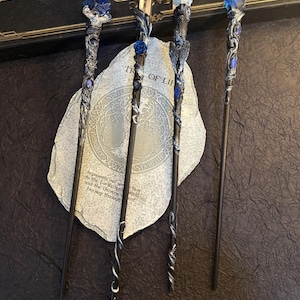 Puede incluir: Cuatro varitas de fantas&iacute;a con dise&ntilde;os intrincados. Cada varita presenta un mango oscuro y una parte superior decorativa con detalles azules o transparentes en forma de cristal. Las varitas se exhiben sobre una superficie similar a una piedra con el texto "TREE OF LIFE" y "HUEHANDIWORK".