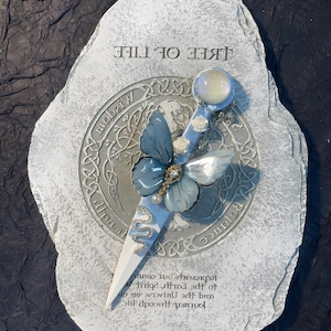 May include: A white stone with a gray and blue butterfly design and the words "Tree of Life" written on it. The stone also has a silver circle with Celtic knotwork and the words "Balance" and "Wisdom" written on it.