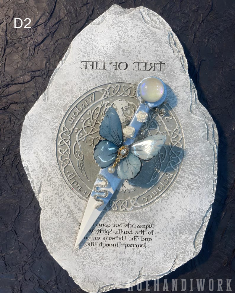 May include: A white stone with a gray and blue butterfly design and the words "Tree of Life" written on it. The stone also has a silver circle with Celtic knotwork and the words "Balance" and "Wisdom" written on it.