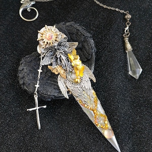 Könnte beinhalten: Ein silberner und goldener Anhänger mit einem Dolch mit Engelsflügeln, einem Schmetterling und einer Sonne. Der Anhänger ist an einer silbernen Kette mit einem Kreuzanhänger und einem Kristallpendel befestigt.