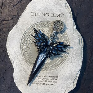 May include: A silver and black metal pendant with a pentagram charm and a blue and black beaded butterfly design. The pendant is on a gray stone with the text "Tree of Life" and "Journey through life and the universe to spirit, earth, and  represents your own",