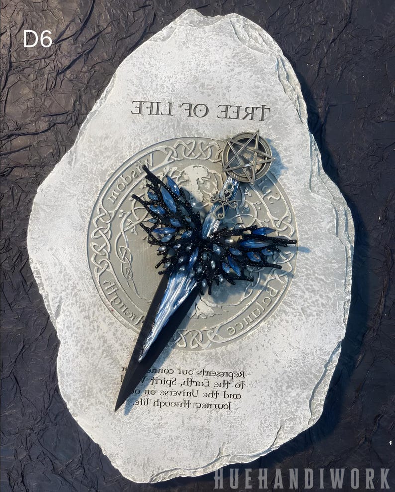 May include: A silver and black metal pendant with a pentagram charm and a blue and black beaded butterfly design. The pendant is on a gray stone with the text "Tree of Life" and "Journey through life and the universe to spirit, earth, and  represents your own",
