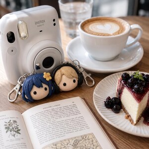 May include: A white Instax mini 11 camera, two small character keychains with blue and blonde hair, an open book with English text, a cup of latte with heart-shaped foam, and a slice of blueberry cheesecake on a white ridged plate.