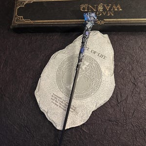 Puede incluir: Una varita m&aacute;gica negra con un mango detallado con detalles azules y una parte superior de cristal azul. La varita descansa sobre una piedra gris con las palabras "TREE OF LIFE" y otro texto. Una caja negra con "MAG WAND" est&aacute; en el fondo.