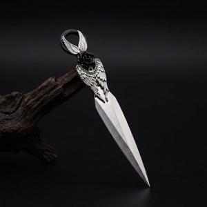May include: A silver dagger with an angel wing design on the handle. The handle features a black rose and a loop for hanging. The blade is sharp and pointed. The dagger is displayed against a dark background.