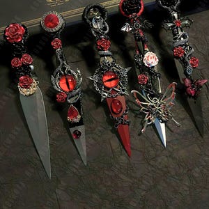 May include: Five decorative daggers with black handles and red rose accents. The daggers have different designs, including a snake, a skull, a butterfly, and a cross. The daggers are arranged on a dark surface with the text "MAGIC WAND" visible in the background.