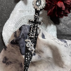 May include: A black dagger with a silver cross and a rhinestone-encrusted snake design. The dagger has a silver-toned, ornate handle with a large, clear rhinestone.