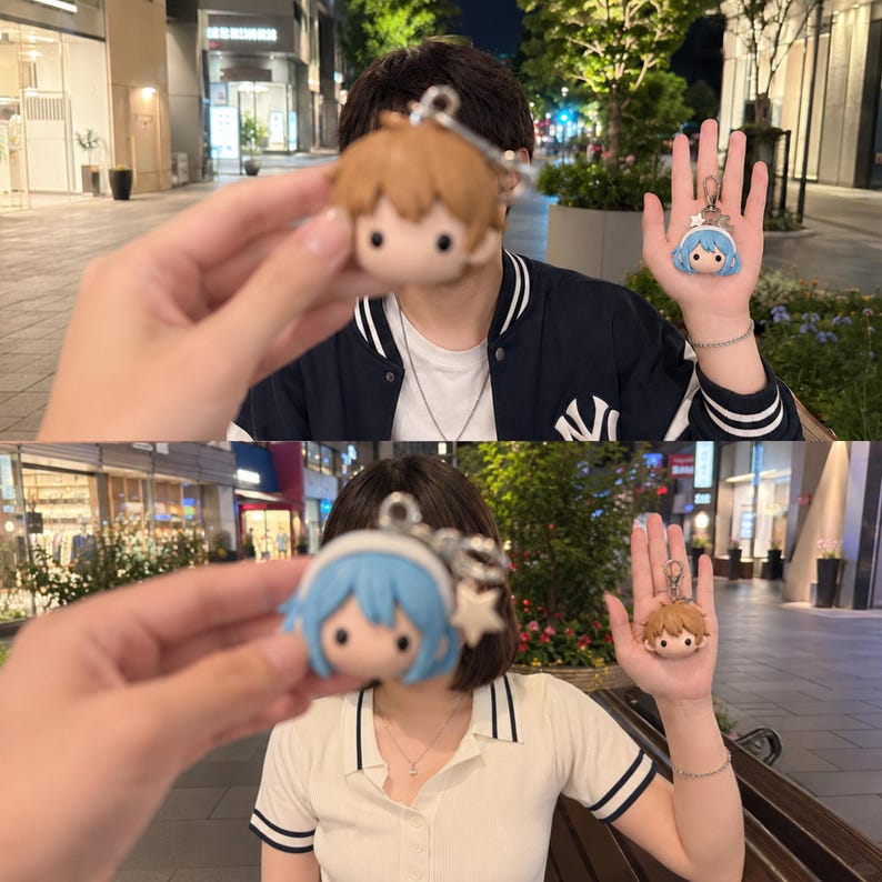 May include: Two small character keychains featuring stylized heads. One keychain has brown hair, and the other has light blue hair with a white headband and a small star charm. Both keychains include a silver metal ring and a lobster clasp attachment.