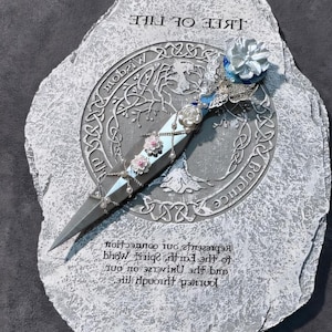 May include: A silver and blue decorative dagger with a floral design, resting on a gray stone with the words "Tree of Life" and a Celtic knot design. The stone also has the text "Journey through life, and the universe on your own. The spirit of the earth, how we connect our present to our connection."