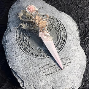 Könnte beinhalten: Ein weißer und grauer Stein mit einem dekorativen Design und den Wörtern "Balance", "Weisheit", "Stärke" und "Leben" um den Rand herum. Ein rosafarbenes und weißes dekoratives Dolch mit einem Schmetterlingsdesign und rosa Blumen liegt auf dem Stein. Der Stein trägt den Text "Stellt unsere Verbindung zur Erde und zum Universum dar. Reise durchs Leben." ein.