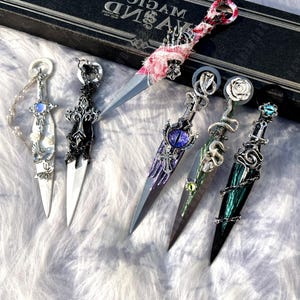May include: An assortment of decorative daggers with ornate handles. The daggers feature various designs, including a skeleton hand, a snake, a rose, and a gemstone. The blades are silver, and the handles are adorned with intricate details and embellishments. A black box with the text "MAGIC" is in the background.