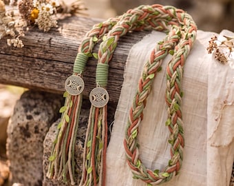 May include: A braided cord made of sage green, terracotta, and cream-colored fabric strands interwoven with small green leaves. The ends are secured with green thread and feature circular silver-toned metal charms depicting a triple moon symbol.