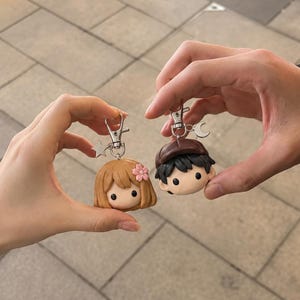 May include: Two character keychains featuring sculpted heads. One has light brown hair with a small pink flower and a silver star charm. The other has dark hair, a brown cap, and a silver crescent moon charm. Both have silver lobster clasps.