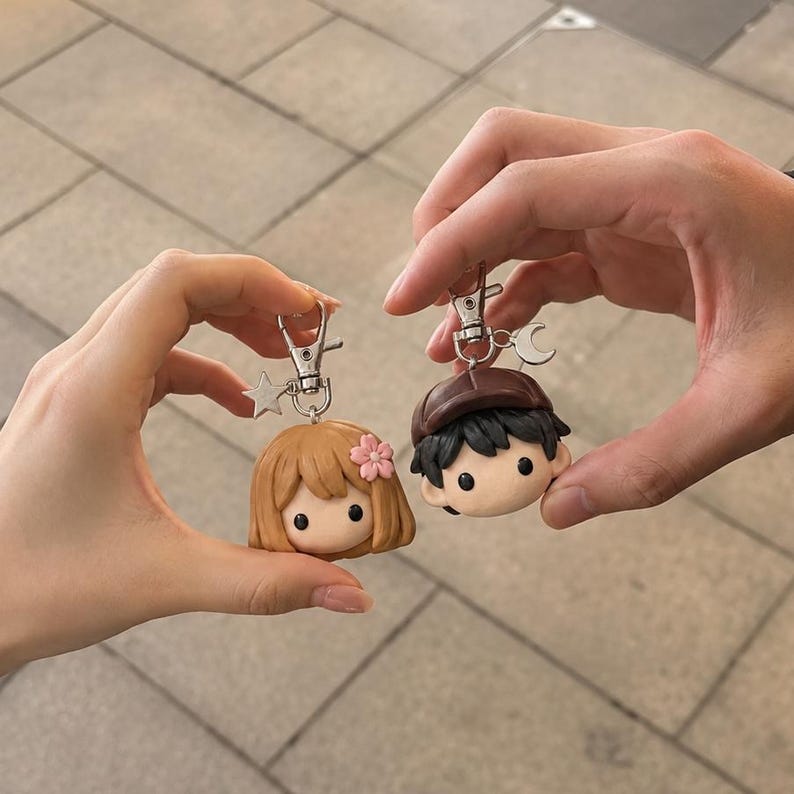 May include: Two character keychains featuring sculpted heads. One has light brown hair with a small pink flower and a silver star charm. The other has dark hair, a brown cap, and a silver crescent moon charm. Both have silver lobster clasps.