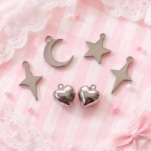 May include: A collection of five silver-toned metal charms featuring a crescent moon, a five-pointed star, two four-pointed star shapes, and two puffed heart charms. Each charm includes a small circular loop at the top for attachment purposes.