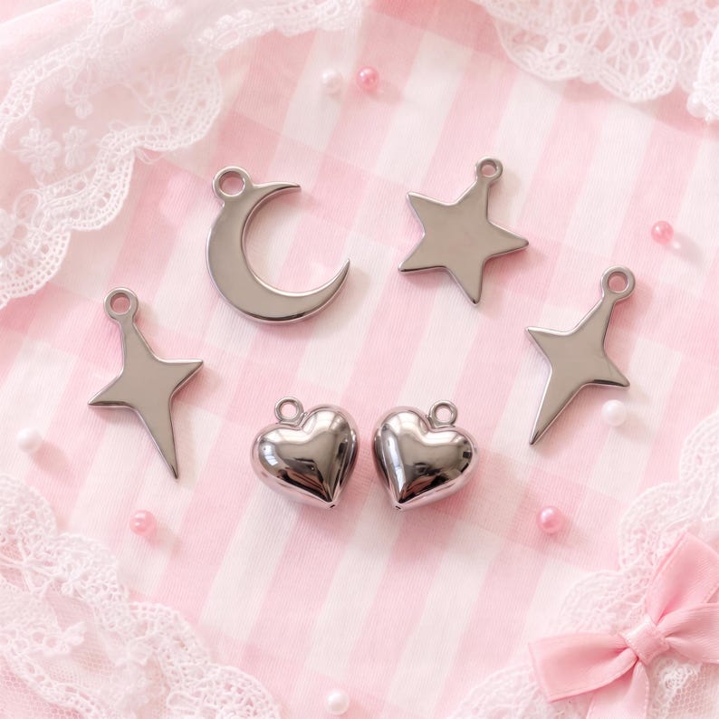 May include: A collection of five silver-toned metal charms featuring a crescent moon, a five-pointed star, two four-pointed star shapes, and two puffed heart charms. Each charm includes a small circular loop at the top for attachment purposes.