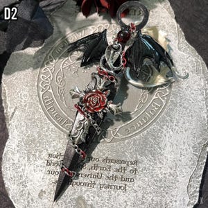 May include: A silver and black pendant featuring a stylized sword with a red rose, a chain, and a black bat wing. The pendant is on a gray background with the words "Journey through and the Universe, represents our Earth, sp", "Balance", and "Wisdom" printed on it.