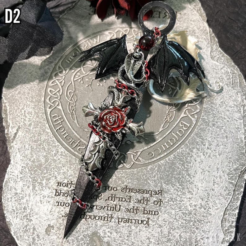 May include: A silver and black pendant featuring a stylized sword with a red rose, a chain, and a black bat wing. The pendant is on a gray background with the words "Journey through and the Universe, represents our Earth, sp", "Balance", and "Wisdom" printed on it.