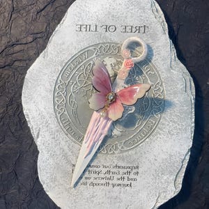 May include: A white ceramic stone with the words "Tree of Life" inscribed on it. A pink and white butterfly with a pearl embellishment is attached to the stone. The butterfly is holding a white and pink wand with a ring at the top.