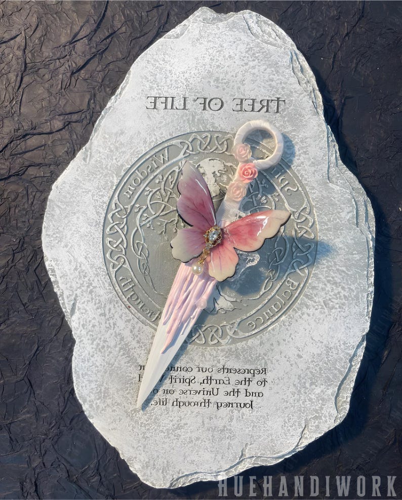 May include: A white ceramic stone with the words "Tree of Life" inscribed on it. A pink and white butterfly with a pearl embellishment is attached to the stone. The butterfly is holding a white and pink wand with a ring at the top.