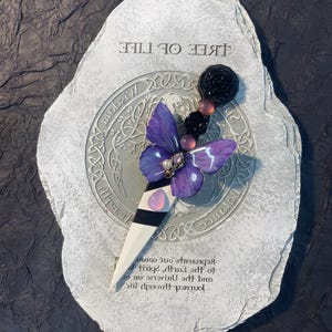 May include: A purple butterfly with a pearl and gold embellishment is attached to a white and black dagger. The dagger is resting on a gray stone with the words "Tree of Life" and a Celtic knot design.