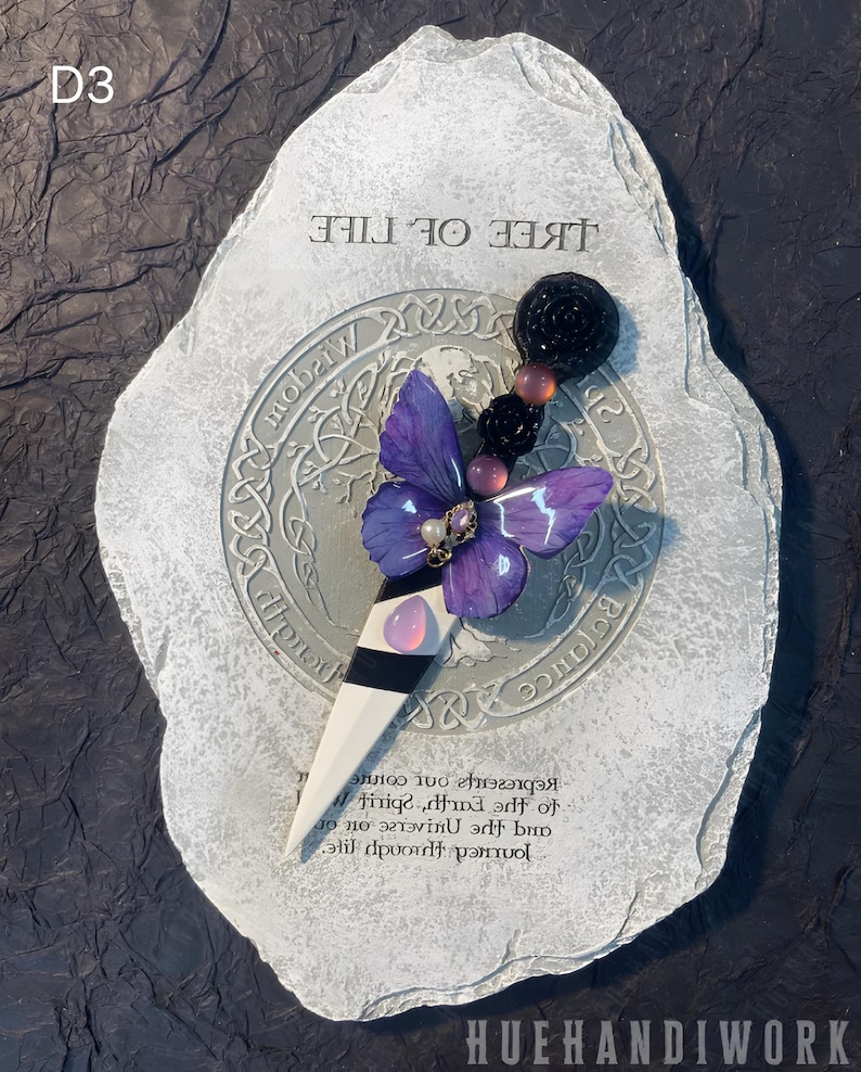 May include: A purple butterfly with a pearl and gold embellishment is attached to a white and black dagger. The dagger is resting on a gray stone with the words "Tree of Life" and a Celtic knot design.