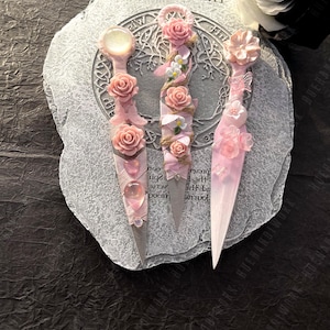 Op de afbeelding: Drie decoratieve roze nail art tools met bloemversieringen. Elke tool heeft een ander ontwerp, waaronder rozen en andere kleine bloemen. De tools worden weergegeven op een getextureerd, lichtgrijs oppervlak.