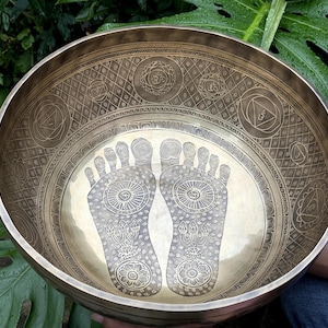 May include: A large, golden singing bowl with intricate engravings. The bowl features two large, detailed footprints in the centre, surrounded by a circular pattern of smaller symbols. The bowl is held by a hand, with green foliage in the background.