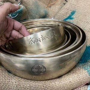 May include: A set of seven nested singing bowls, made of brass, with a Tibetan mantra inscribed on the largest bowl. The bowls are arranged in a stack, with the largest bowl at the bottom and the smallest bowl at the top.