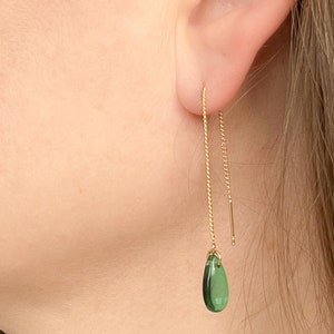 Może przedstawiać: Zbliżenie złotego kolczyka łańcuszkowego z zielonym kamieniem szlachetnym w kształcie łzy. Kolczyk jest włożony w ucho. Tło to skóra i włosy osoby.
