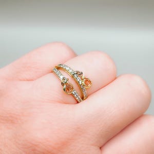 May include: A gold ring with two dangling stones. The ring is set with small clear stones and the dangling stones are a light orange color.