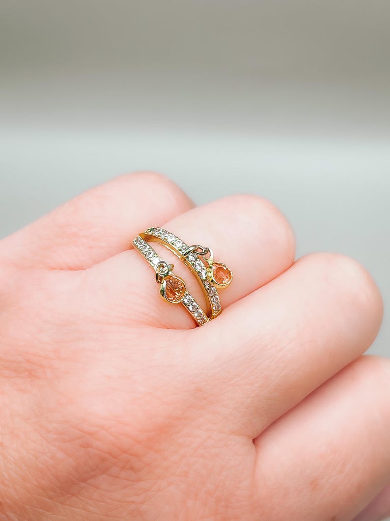 May include: A gold ring with two dangling stones. The ring is set with small clear stones and the dangling stones are a light orange color.