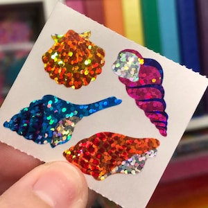 May include: A sheet of colorful iridescent stickers featuring four different seashells. The stickers are on a white background and have a glittery finish.