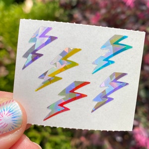 May include: A sheet of six holographic lightning bolt stickers in various colors, including purple, yellow, and red. The stickers have a geometric design and are displayed against a blurred green and pink background.