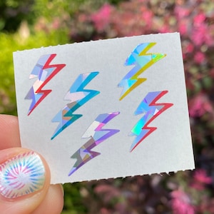May include: A small white rectangle with six holographic lightning bolt stickers in various colors. The lightning bolts have red, blue, yellow, and purple accents. The stickers are held up by a person's hand with a tie-dye nail design.