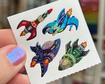 May include: A small, white square featuring five colorful rocket ship illustrations. The rockets are red, blue, green, and purple with glittery accents and flames. The image is held by a person.