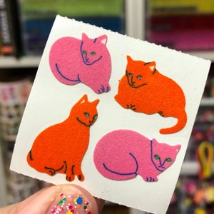 May include: Four colorful cat stickers on a white sheet of paper. Two of the cats are pink and two are orange. The cats are all in different poses.