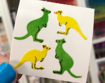 May include: Four colorful felt kangaroo stickers on a white sheet of paper. Two kangaroos are green and two are yellow.