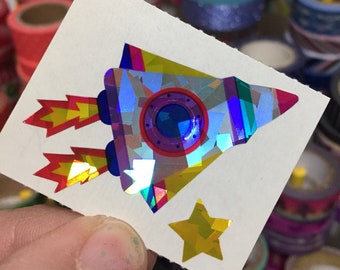 May include: A white sticker sheet with a holographic rocket ship and a gold star. The rocket ship is blue, purple, and yellow with a red window. The sticker sheet is surrounded by rolls of colorful washi tape.