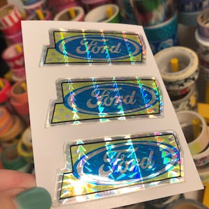 May include: Three holographic stickers with the word "Ford" in a blue oval on a yellow background. The stickers are on a white sheet of paper.