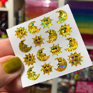 May include: A sheet of gold holographic stickers featuring suns and moons with smiling faces. The stickers are on a white background.