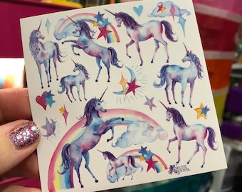 May include: A sheet of unicorn stickers featuring watercolor illustrations of unicorns, rainbows, stars, and clouds. The unicorns are depicted in shades of blue, purple, and pink. The stickers have a whimsical and magical theme.