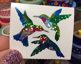 May include: A white sticker featuring three iridescent hummingbird designs. The hummingbirds have blue, green, and red accents, with a shimmering, glittery effect. The sticker is held in front of a background of colorful washi tape rolls.