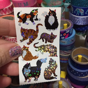 May include: A sheet of holographic cat stickers with a variety of poses and colors. The stickers are on a white background and have a glittery finish.