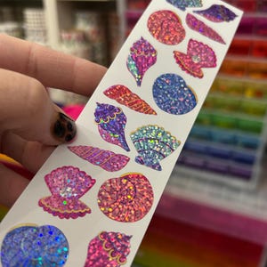 May include: A strip of seashell stickers in various shapes and colors, including pink, blue, and iridescent glitter. The stickers are held against a blurred background of shelves and colorful items.