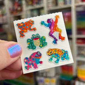 May include: A sheet of five colorful frog stickers. The frogs are in various poses and feature vibrant patterns in orange, blue, pink, green, and yellow. The stickers have a glossy finish and are on a white background.